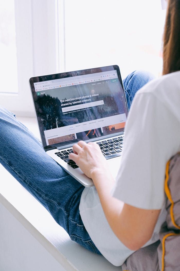 Young woman in casual jeans working on a laptop indoors by a window, showcasing remote work.